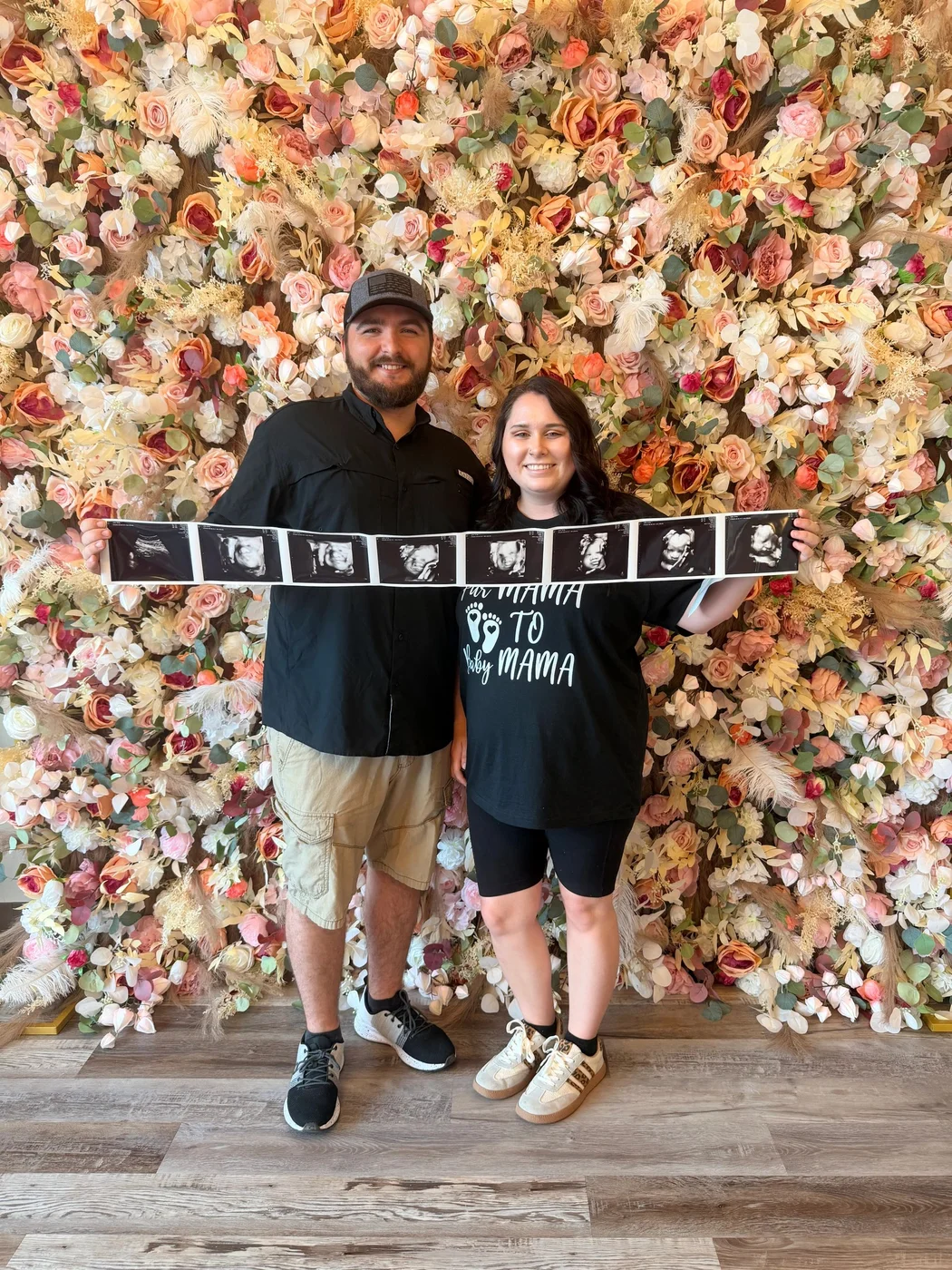 Two guests holding ultrasound prints in front of a floral studio wall